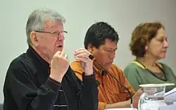 Two men and a woman are sitting behind a table at a conference. One of the men is giving a speech.
