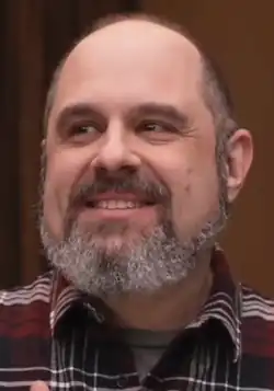 A 51-year-old man with a grey beard smiling to the left of the camera.