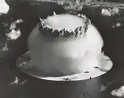 Aerial photo of nuclear explosion rising from lagoon. Hemispherical condensation cloud on the surface is 1 mile (1.6&nbsp;km) in diameter. In comparison, Navy ships in the foreground look like bathtub toys.