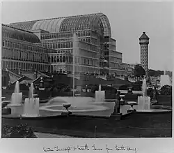 A black and white photo of a massive glass building with a tower to the right and fountains in front. Below, in cursive script, is a caption that reads "Centre transept & north tower from South Wing"