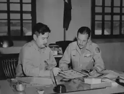 A black and white photograph of two seated uniformed men inspection documents while smoking a cigar and a pipe