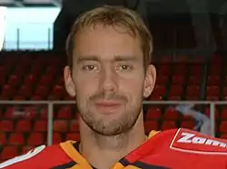 An ice hockey players face and shoulders. He has short brown hair and is not wearing a helmet.