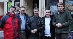 Five middle-aged, white men standing should-to-shoulder in front of a restaurant and smiling at the camera. They wear casual clothing and winter jackets.