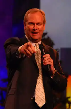 Dan Clark makes a speech. He holds a microphone and is pointing toward the crowd.