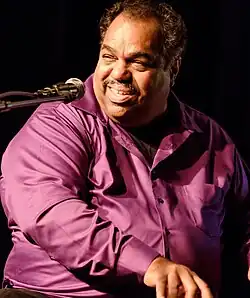 Daryl Davis wearing a purple button-down shirt, playing a piano, turned right-of-camera, towards a microphone