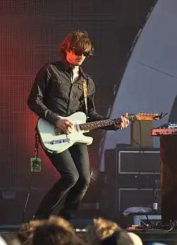 Middleton is playing his guitar with his left hand on the fret board and right hand on the strings. His knees are flexed, he wears dark glasses, dark pants and a dark shirt. His stage pass dangles from the end of the guitar. His hair is brown and hangs partly into his eyes. He wears a wide ring on his right hand's fourth finger. There is band equipment nearby.