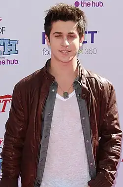 A mid shot of a man with spiked, light brown hair, wearing a brown, leather jacket. He is posing at a press event and smiling, looking directly towards the camera. Behind him are hoarding signs with names of commercial sponsors.