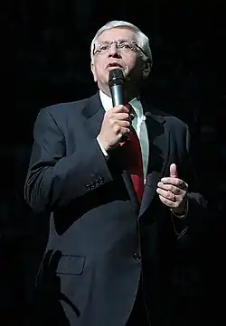 A man in his 50s with white hair speaks into a microphone. He wears a gray suit, red tie, and glasses.