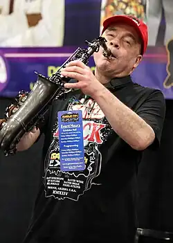 David Wise, a British man wearing a red cap and black shirt, plays a saxophone
