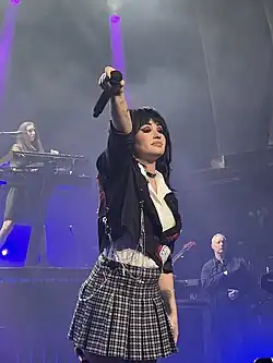 Demi Lovato holding a microphone and wearing a white shirt, black jacket, and checkered skirt.