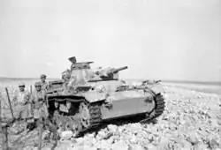 A tank lies crippled in the desert with soldiers standing around it