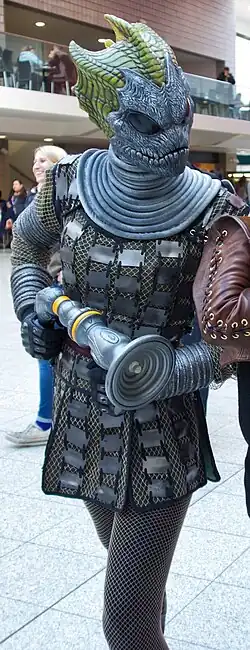 A lizard-like humanoid, wearing netted clothing and a grey mask on its head. Its skin is green and lizard-like, and its mask bears bulbous black eyes.