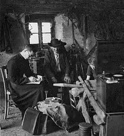Three white people seated in a barn in 1939; a man in priest's clothing, a farmer at center, and a woman with her head down, writing; a recording device is propped on an overturned wheelbarrow in front of the group.
