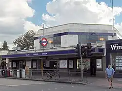 A grey building with a blue sign reading "EALING COMMON STATION" in white letters and two green trees in the background all under a blue sky