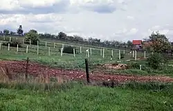 A hillside with multiple barbed-wire fences running parallel to each other, with fruit trees, a barn and a watchtower in the background.
