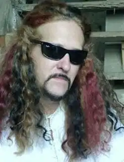 Three-quarter profile colour publicity shot of a man wearing sunglasses, with serious expression, minimal moustache-beard combo, and cascades of long, wavy brown, red, and black hair.