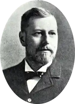 A black and white portrait of a bearded man, wearing a suit with a bowtie