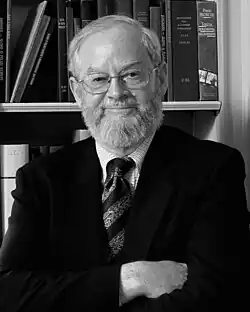 A black and white image of a balding man in a suit with a full beard and glasses standing in front of a bookshelf with his arms crossed smiling
