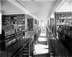 The second floor of a library with thin rails; the bottom floor is visible and in the distance is a spiral staircase