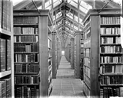 A massive row of bookcases terminating at a diamond-patterned window while a pointed roof above is made mostly of windows with sunlight beaming through