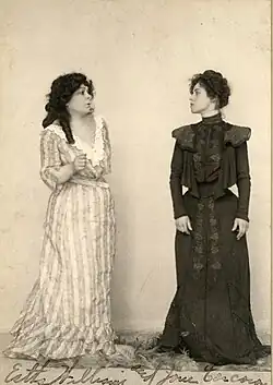 Two white women, standing, wearing long dresses, facing each other.