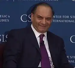 A man with short black hair wearing a suit and standing in front of a blue background
