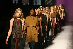 A line of models on a catwalk, each wearing a unique outfit with a black and brown color scheme