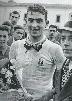 A man wearing a white jersey with goggles around is neck holding a bunch of flowers.