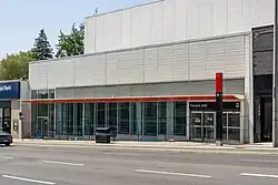 Photo of a light rail station entrance under construction which appears largely completed. It is a white building with glass panelling and an orange accent.