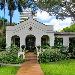 A brick walkway leads from a lush garden to a gray stucco building.