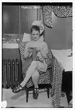 A young white woman wearing a theatrical costume, seated indoors, knitting