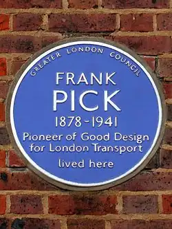 A circular blue ceramic plaque with white raise lettering fixed to a brick wall bears the text "FRANK PICK, 1878–1941 Pioneer of Good Design for London Transport lived here"