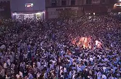 A large gathering of people on a street with a bonfire in the right side of the image.