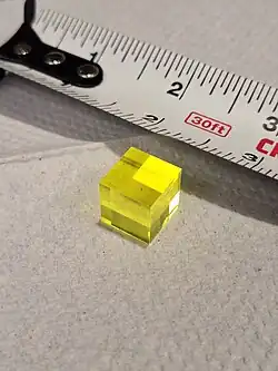 A bright yellow, transparent, cubic crystal rests on a paper towel. A tape measure in the background gives a size comparison, indicating that the cube is 5cm on each side length.