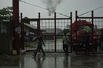 A picture of gates of the Port Harcourt Refining Company Ltd. with an oil refinery's fire seen in the background in rainy weather.