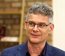 close-up of Geoffrey Miller wearing a dark purple shirt and a dark blue blazer, smiling and looking right of camera