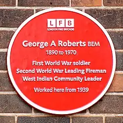 Circular red plaque, with six lines of text reading "George A Roberts BEM / 1890 to 1970 / First World War Soldier / Second World War Leading Fireman / West Indian Community Leader / Worked here from 1939". Above those, the text and logo "London Fire Brigade".