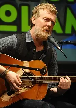 close-up image of Glen Hansard wearing dark jeans, a blue checkered shirt, and grey vest, seated, playing acoustic guitar and singing into a microphone onstage