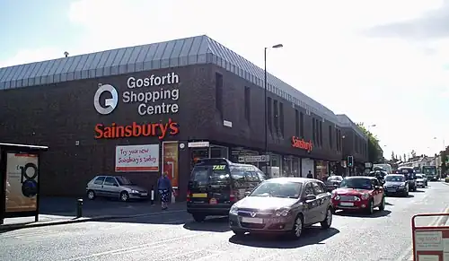 A 1970s monolithic shopping centre, mostly grey/brown in colour. In the foreground the busy high street with a number of travelling cars.