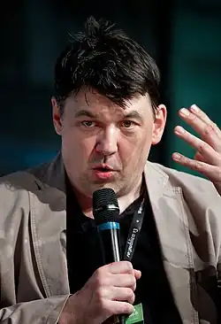 A photograph of Linehan. Linehan is talking into a handheld microphone while making a gesture with his other hand. He is wearing a jacket, shirt, and lanyard.