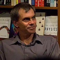 Graham Oppy wearing a grey shirt with a lapel microphone, bookcases in background, looking right of camera with slight smile