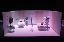 A hand-made scale model of a hospital room separated by a curtain, with grapes standing in for patients. On the left of the curtain, there are two "patients" wearing surgical masks, one in a bed connected to an electrocardiogram and one sitting down on a chair besides it. On the right, there is a surgical bed where the da Vinci Surgical System performs surgery.