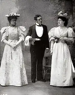 Butler between two young women, all dark-haired and all standing, in a garden setting; the women, both in hats, regard each other angrily