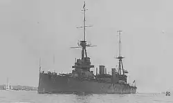 Photograph of a large warship. Along the bottom edge of the photograph, the phrases "HMAS AUSTRALIA" and "BUILT FOR THE AUSTRALIAN NAVY" are written in white. A sailboat and distant land can be seen in the left part of the photo.