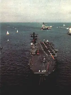 Colour photo of a small aircraft carrier, taken from an elevated position. The carrier is sitting bow on, with one of her anchor chains deployed.