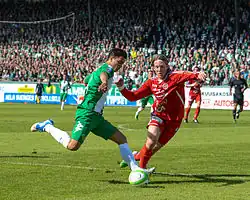 A soccer player dressed in green and white runs with the ball. A player in red and white tries to take the ball. In the background there are stands filled with Hammarby fans.