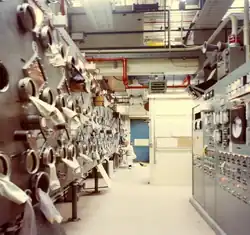 A color photograph of a man, seen on the bottom left of the image, operating a complex machine which has glass openings and is covered behind a red metallic front