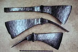 The top tool has a long pick with a verticla chisel-like edge, parallel to the broad axe head on the other end, the top of which is in line with the straight, untapering "pick", the bottome edge of which flares in the shape of a pendant equilateral triangle, save that the sharp edge is curved. The lower two tools have blades that are a bit closer to vertical, and narrower transverse azde heads, which are bent down so that they would form an angle of about 60 degrees to the missing wooden shafts.