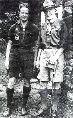 two men outdoors, both wearing Scout uniforms: short sleeve shirt, shorts and knee socks, the older man on the right wearing a campaign hat and holding a cane