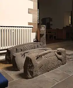 This image shows a collection of early medieval hogback tombstones displayed within a church interior. These distinctive Viking-age monuments are carved from weathered sandstone and exhibit the characteristic house-like form that gives them their name, resembling the shape of Viking longhouses with curved ridged roofs. The hogbacks feature intricate carved decoration including geometric patterns, interlacing designs, and what appears to be representations of roof tiles or 'shingles'. Some examples show carved end-beasts - stylised animal figures at the terminals. The monuments are arranged on the stone floor near church furnishings including wooden chairs and a white radiator. These rare survivals from the Anglo-Scandinavian period (roughly 10th-11th centuries) are primarily found in areas of northern England that experienced Viking settlement, representing some of the earliest forms of commemorative sculpture in English churches.